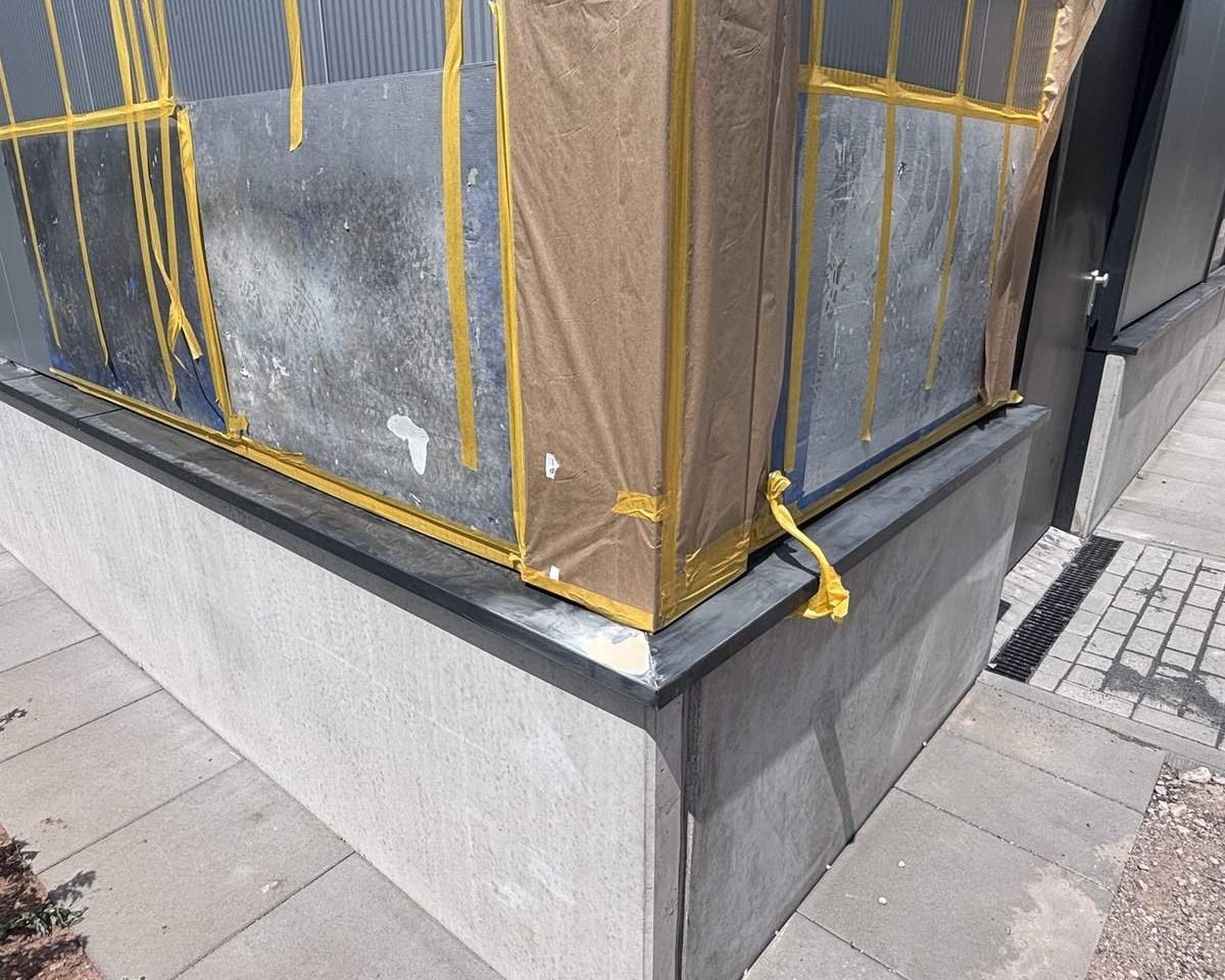 Dieselbe Fassadenecke vor der Reparatur: sichtbare Kratzer und eine Verformung am Tropfblech, Bereich zur Lackierung abgeklebt.